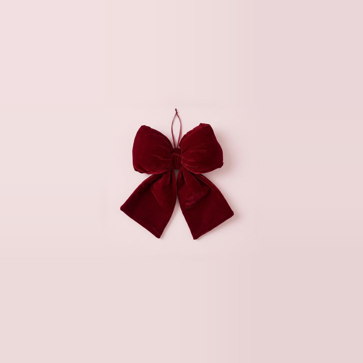 A wine-red velvet Christmas bow with gathered loops, two flared tails, and a hanging ribbon sits centered on a pale pink background.