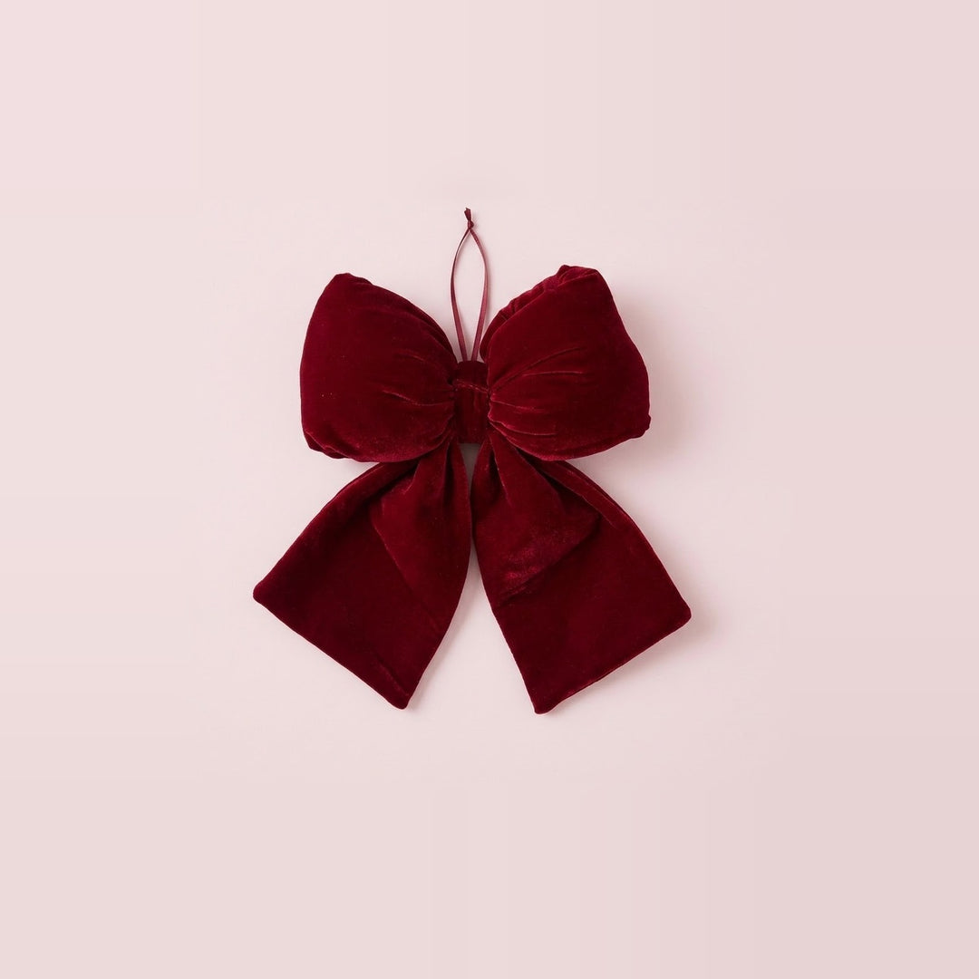 A wine-red velvet Christmas bow with broad tails and a thin hanging loop rests against a pale pink background.