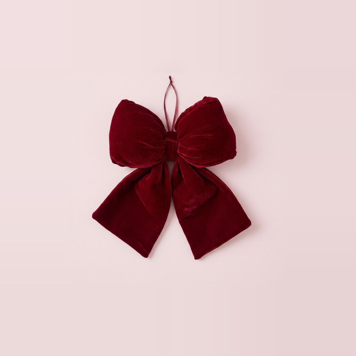 A wine-red velvet Christmas bow with broad tails and a thin hanging loop rests against a pale pink background.