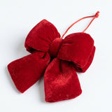 A red velvet Christmas bow lies diagonally on a white surface, showing layered loops, broad tails, and a knotted hanging cord.