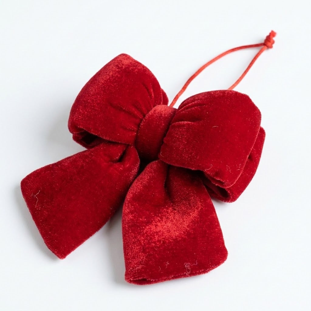A red velvet Christmas bow lies diagonally on a white surface, showing layered loops, broad tails, and a knotted hanging cord.