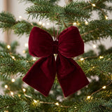 A burgundy velvet bow hangs among green Christmas tree branches surrounded by warm white string lights.