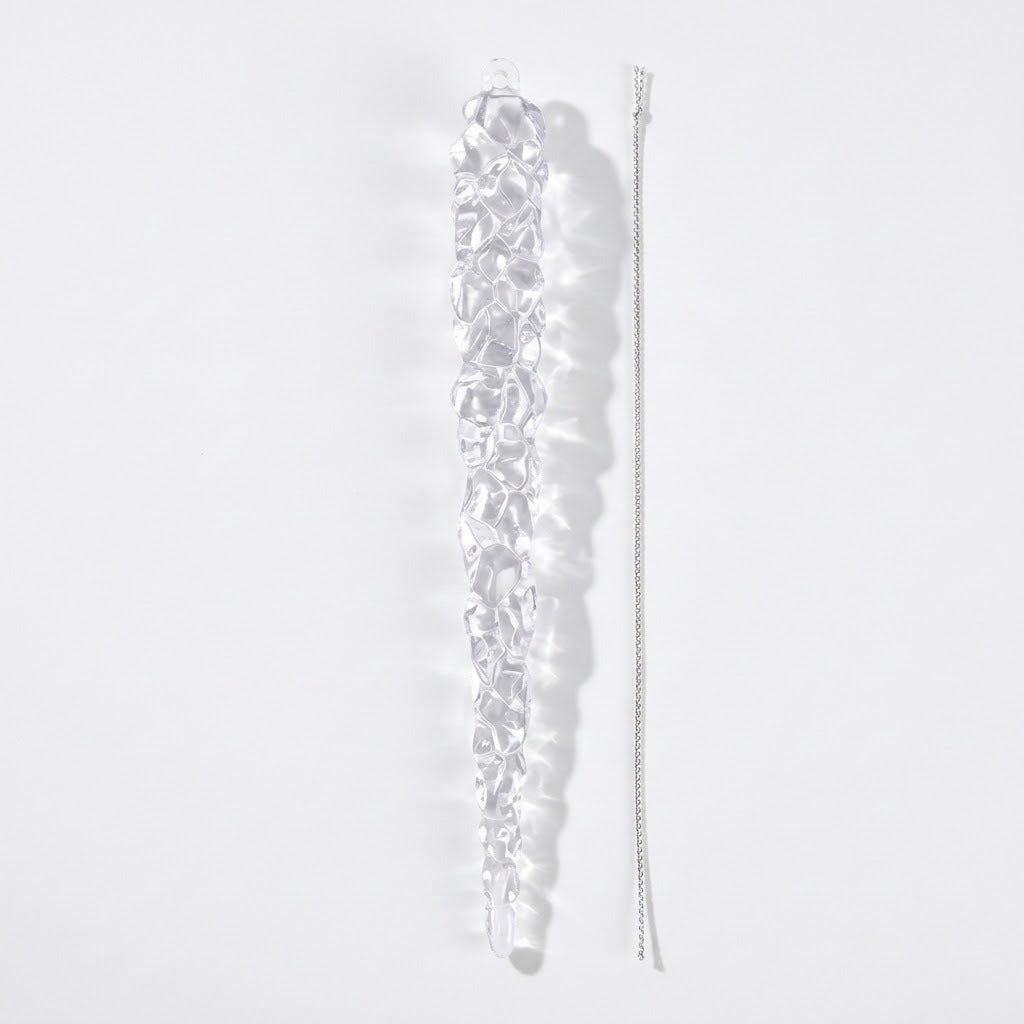 A clear tapered icicle ornament lies beside a separate silver hanging cord on a white background, casting a soft shadow.