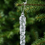 A close-up shows a clear icicle ornament suspended by a silver cord through its circular eyelet on a green tree branch.