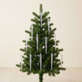 Clear icicle ornaments hang from a small green Christmas tree against a cream wall above a pale wooden floor.