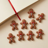 Nine brown resin gingerbread craft embellishments with red bows and pale blue buttons sit beside an open cream book with a red ribbon.