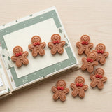 Brown resin gingerbread craft embellishments with red bows and brown buttons decorate a green-bordered scrapbook page and the surrounding wooden surface.