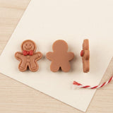 Brown resin gingerbread craft embellishments show their decorated front, plain flat back, and side profile on cream paper beside red-and-white twine.