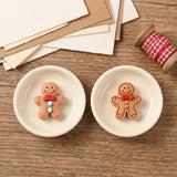 Two brown resin gingerbread craft embellishments sit in cream dishes beside paper and red gingham ribbon on a wooden surface.