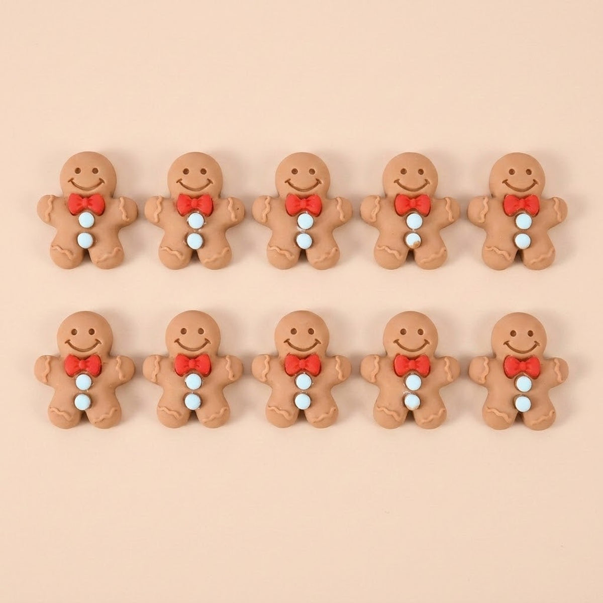 Ten brown resin gingerbread craft embellishments with red bows and pale blue buttons sit in two rows against peach.