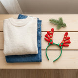 A green Christmas headband with red antlers and gold bells lies beside a cream sweater and blue jeans on pale wooden slats.
