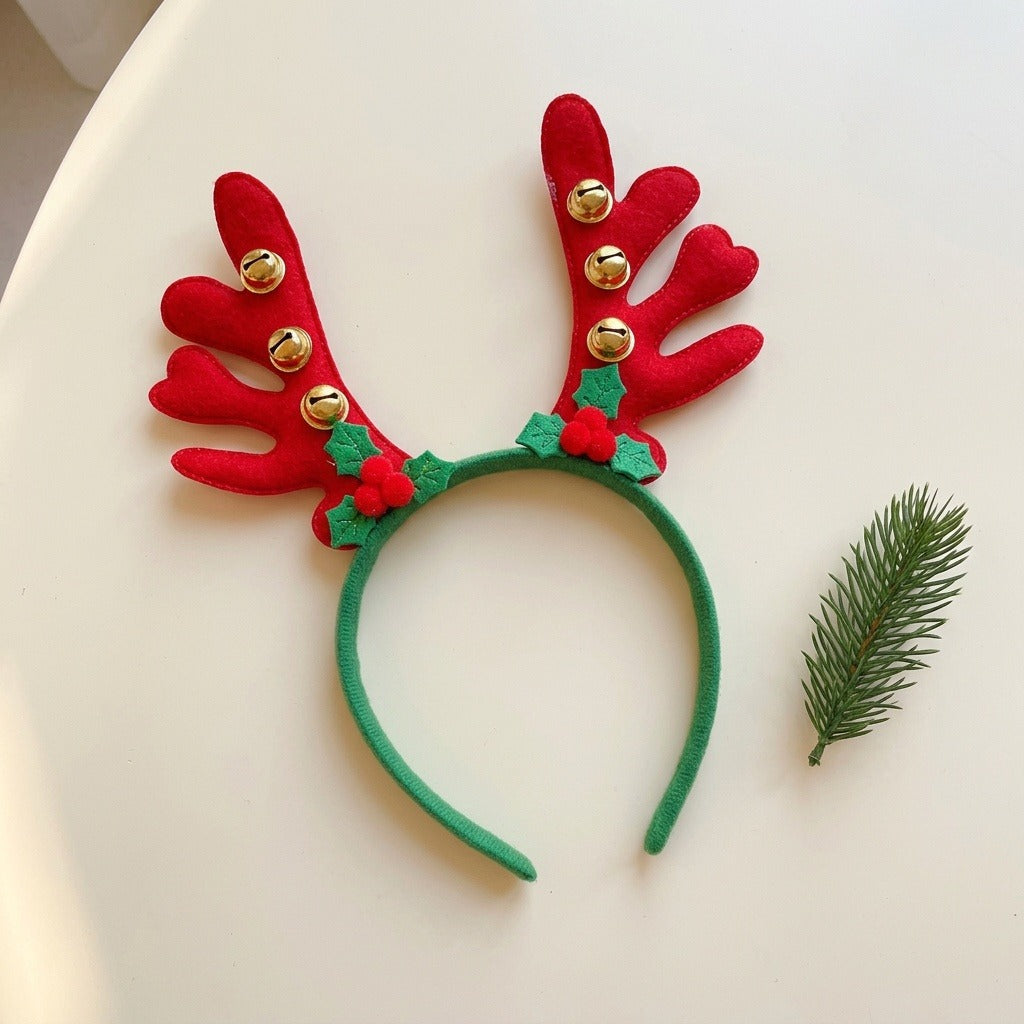 A green fabric Christmas headband with red antlers, six gold bells, and holly rests beside an evergreen sprig on a cream surface.