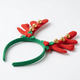 Green fabric Christmas headband with red antlers, six gold bells, holly leaves, and red pom-pom berries lies angled on white.