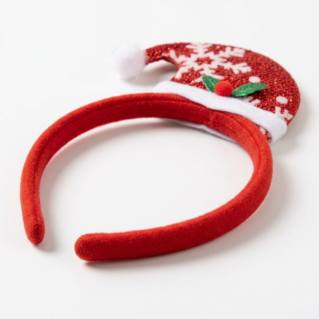 Red fabric Christmas headband with a glittery Santa hat, white snowflakes, fluffy trim, and green holly leaves rests on white.