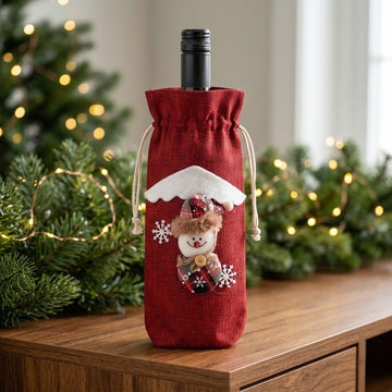 A red woven wine bottle cover with a snowman decoration stands on a wooden cabinet beside evergreen branches and warm Christmas lights.
