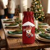 A red cloth wine bottle cover with a snowman decoration stands on a festive wooden dining table with green napkins.