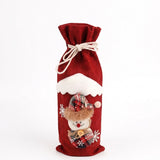 A red woven wine bottle cover with cream drawstrings, white snowflakes, and a plaid snowman decoration stands against a white background.