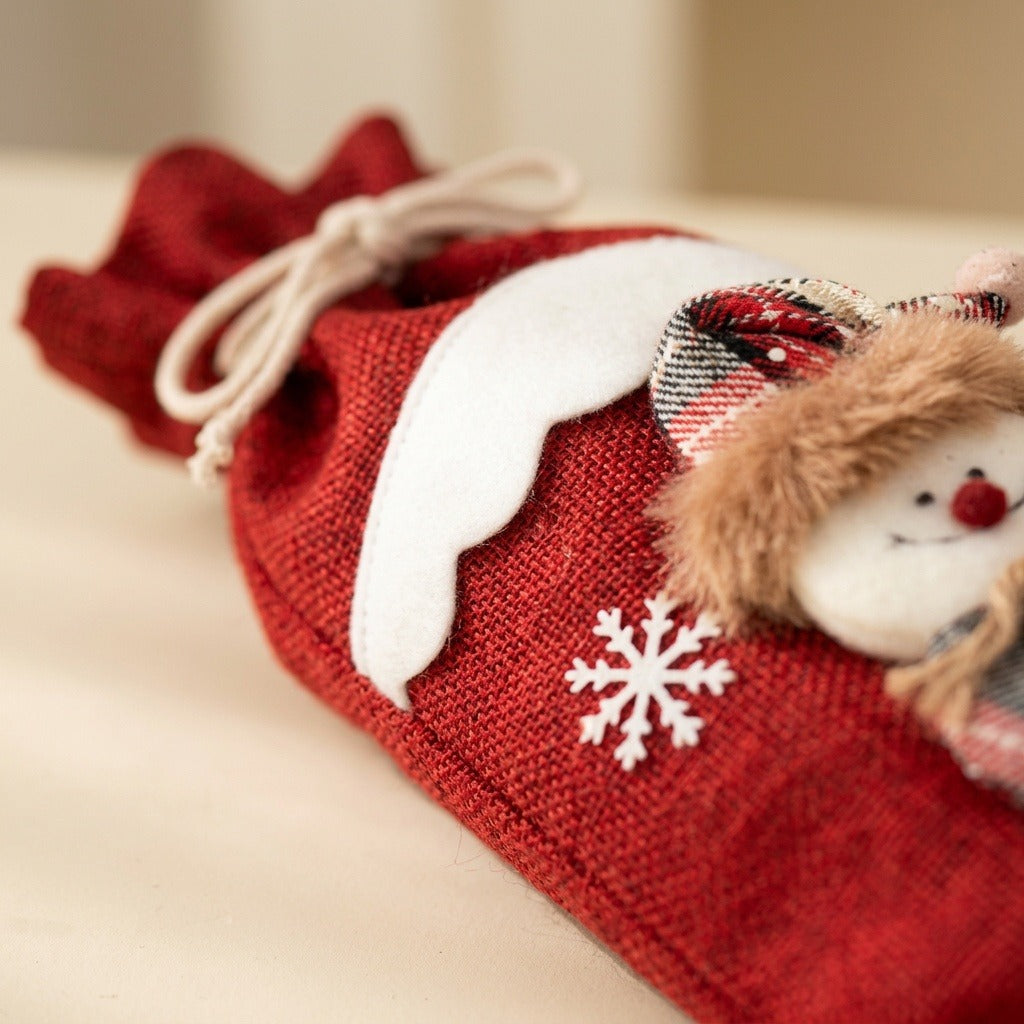 A red woven wine bottle cover lies on a cream surface, showing white scalloped trim, a snowflake, and a plush snowman detail.