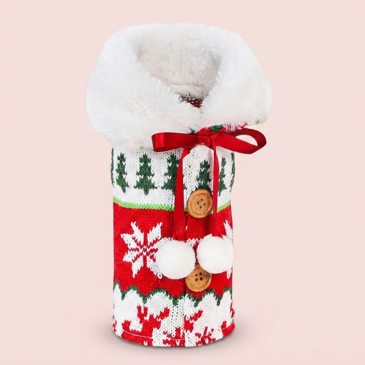 Red, green and white knitted wine bottle cover with fluffy trim, buttons and pom-poms stands against a pale pink background.
