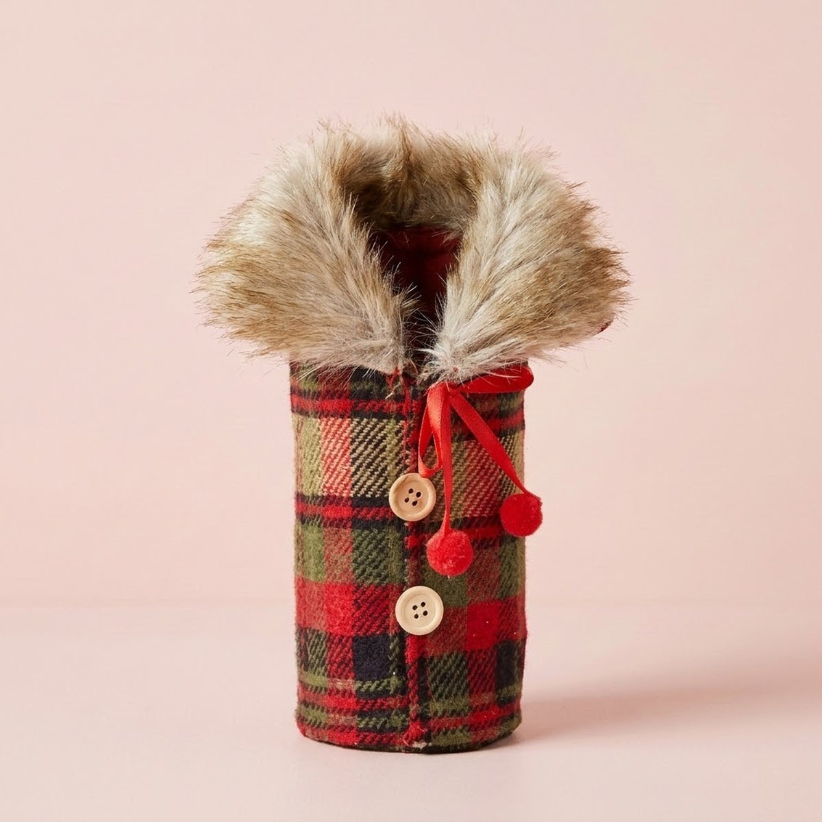 Red and green plaid wine bottle cover has a fluffy tan collar, two beige buttons and red pompom ties against a pale pink background.