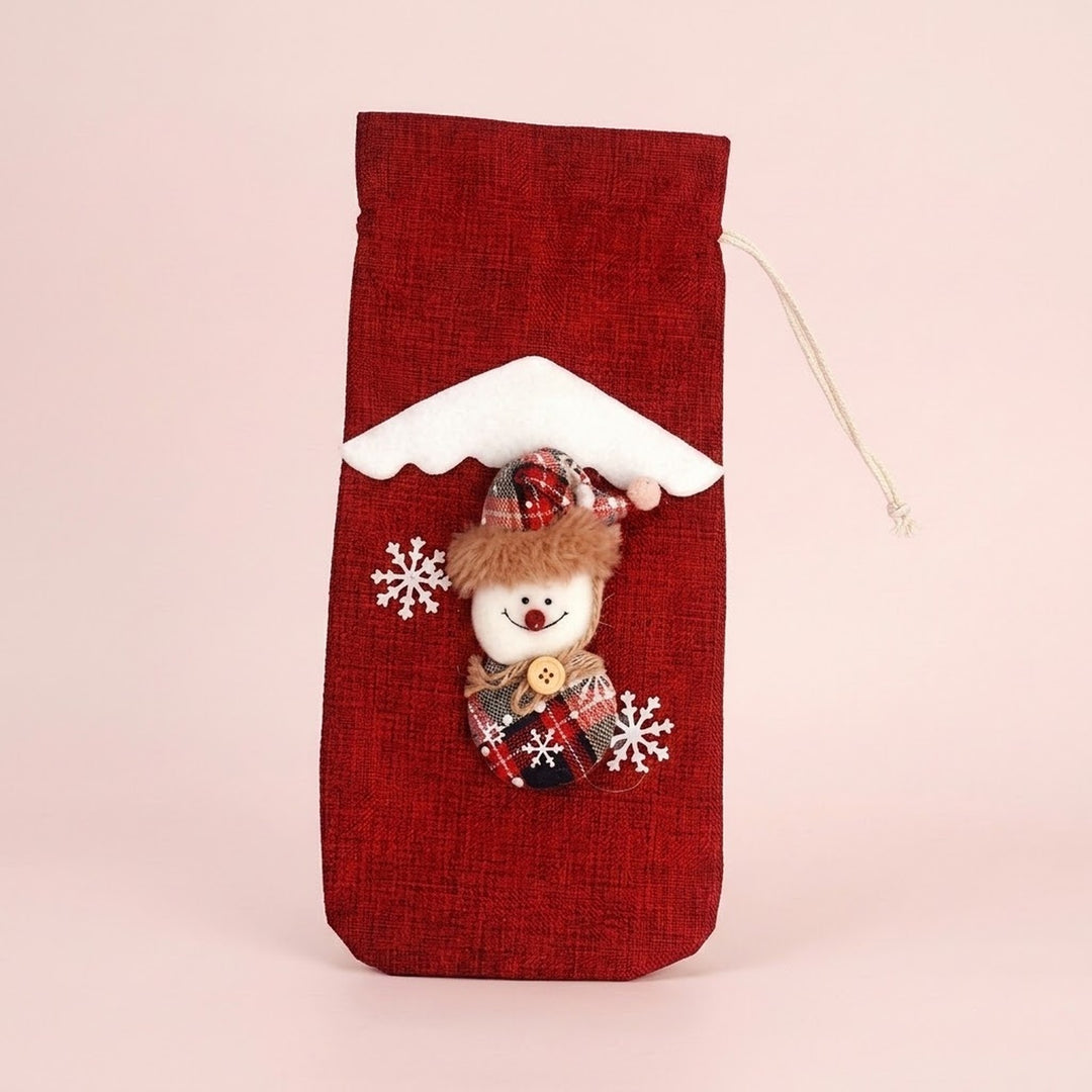 Red drawstring wine bottle cover with a snowman appliqué, white snowflakes and snowy roof trim against a pale pink background.