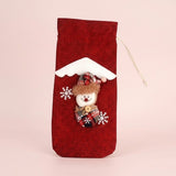 Red drawstring wine bottle cover with a snowman appliqué, white snowflakes and snowy roof trim against a pale pink background.