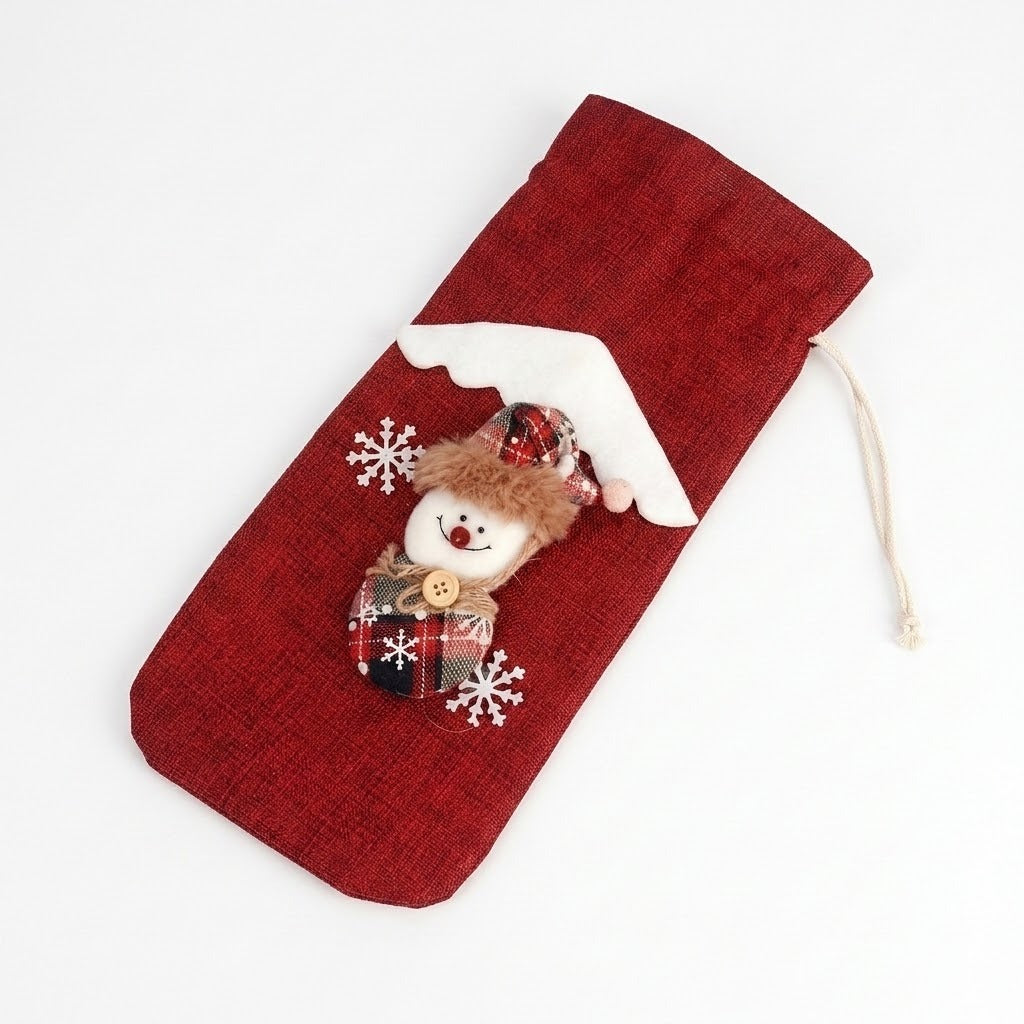 Red woven wine bottle cover lies diagonally on white, decorated with a plaid snowman, white snowflakes and cream drawstrings.