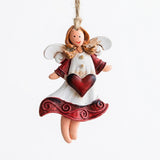 A resin angel Christmas ornament with white wings, golden hair, and a burgundy heart hangs from twine against a white background.