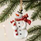 A white resin snowman Christmas ornament with a black hat, red scarf, and candy cane hangs among evergreen branches against a cream background.