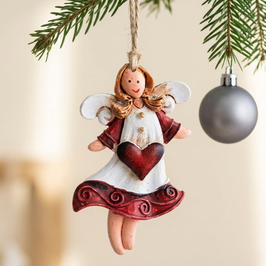 A white and burgundy angel Christmas ornament hangs beneath evergreen branches beside a silver bauble against a beige background.