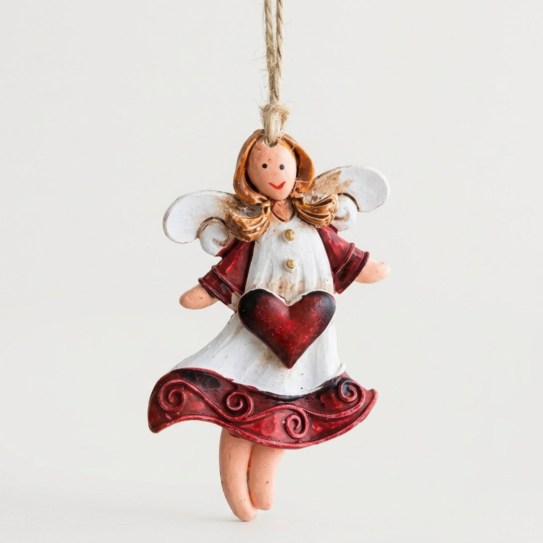 Resin angel Christmas ornament with white wings, burgundy sleeves and a red heart hangs from twine against an off-white background.