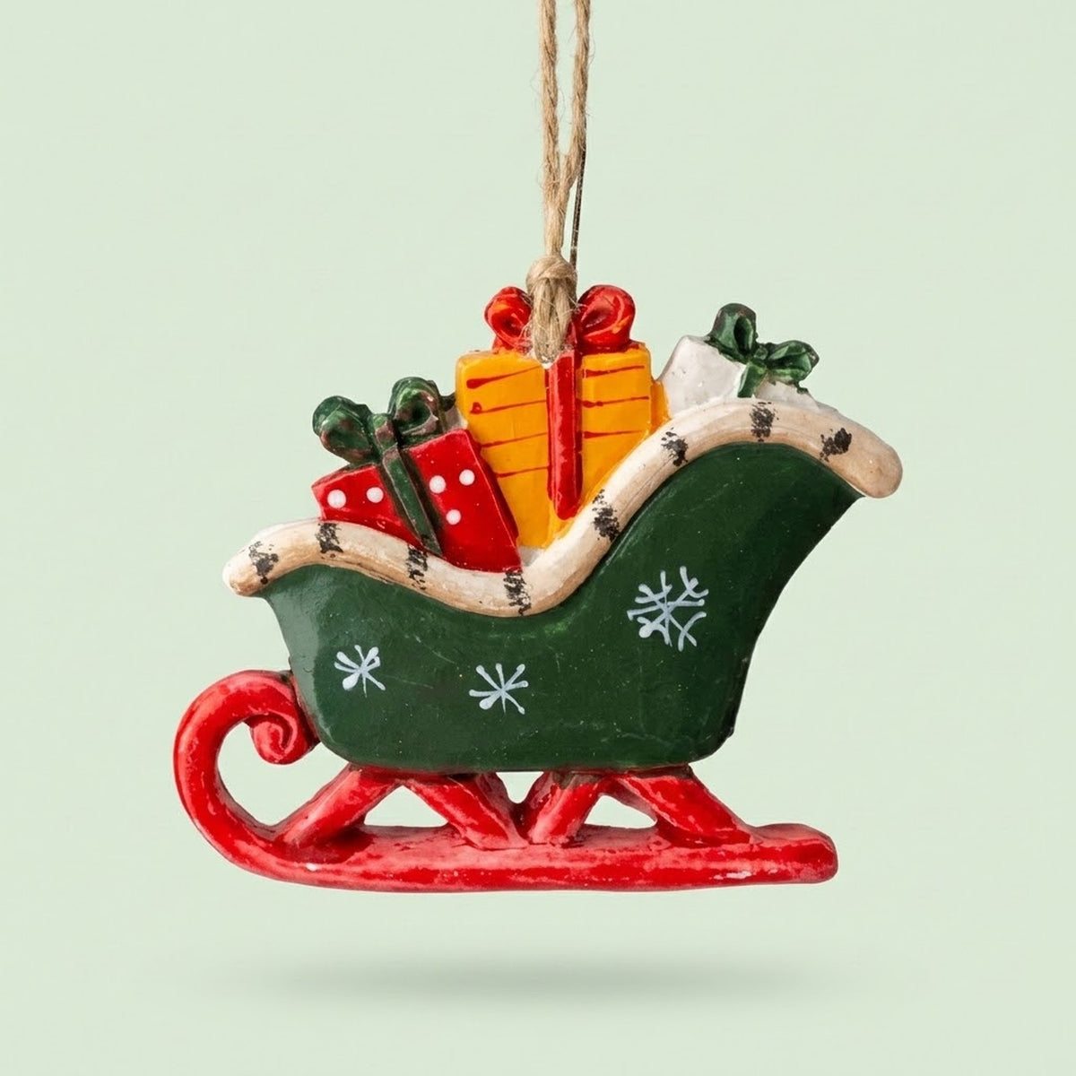 A green sleigh Christmas ornament with white snowflakes, red runners, and colorful gifts hangs by twine against a pale green background.
