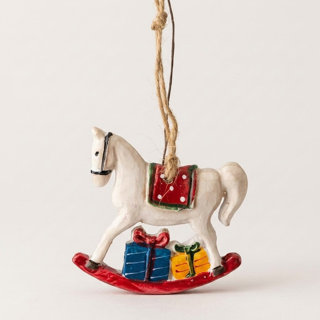A white rocking horse Christmas ornament has a red saddle, blue and yellow gifts, and red rockers against an off-white background.