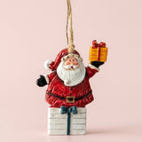 A red resin Santa Christmas ornament holds a yellow gift above a white present with teal ribbon against a pink background.