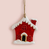 Resin Christmas ornament shaped as a red house with a snowy white roof and black doorway hangs against a pale pink background.