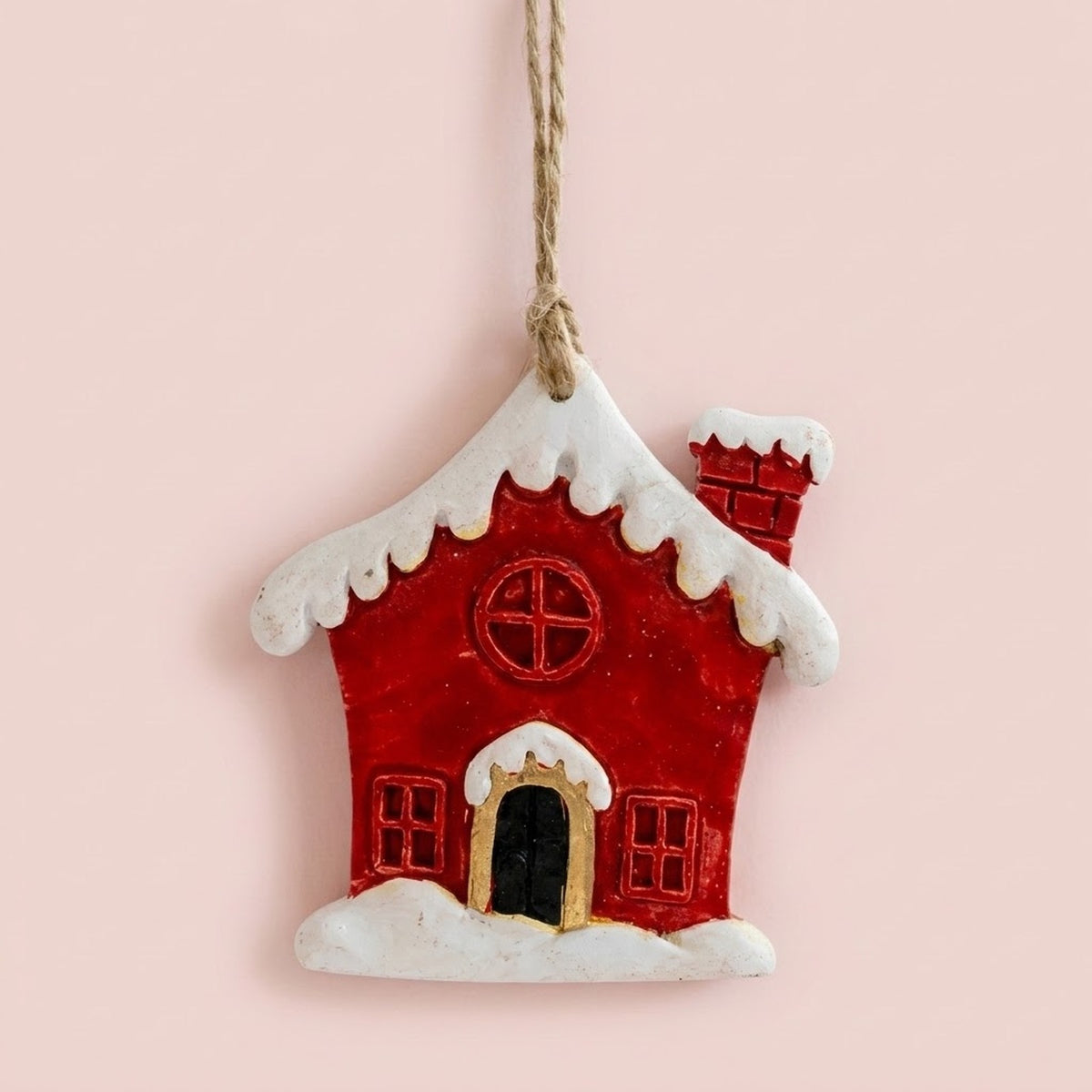 Resin Christmas ornament shaped as a red house with a snowy white roof and black doorway hangs against a pale pink background.