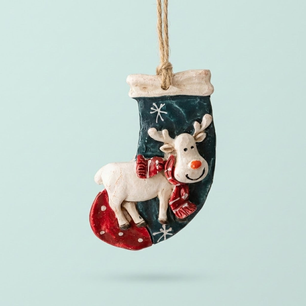 Resin Christmas ornament shaped as a teal stocking with a white reindeer, red scarf and snowflakes hangs against a pale mint background.