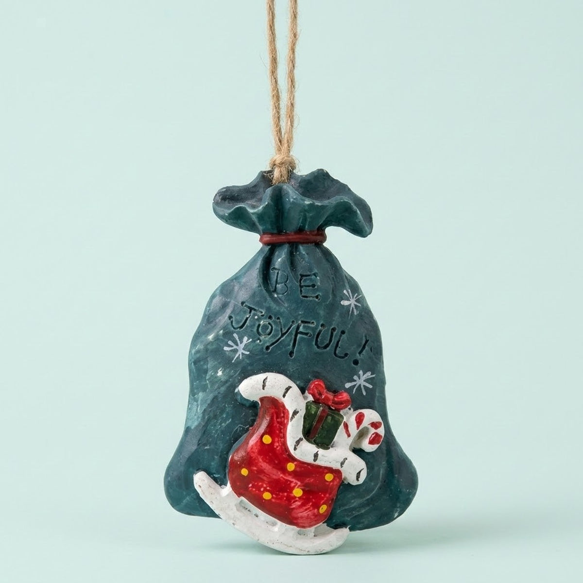 Resin Christmas ornament shaped as a teal gift sack reading “BE JOYFUL!” features a red sleigh and candy cane against a mint background.