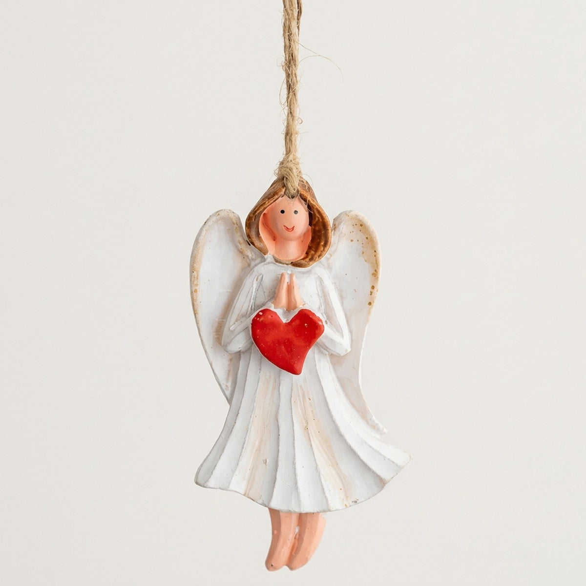 Resin Christmas ornament depicting a white angel with praying hands and a red heart hangs by twine against an off-white background.
