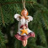A white and burgundy resin angel Christmas ornament holding a yellow star hangs from twine against dense green evergreen branches.