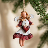 A white and burgundy resin angel Christmas ornament with a red heart hangs beside evergreen needles against a blurred beige background.