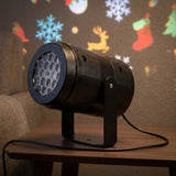 Black Christmas light projector with circular lenses stands on a wooden table beneath colorful snowflake, reindeer, and stocking projections.