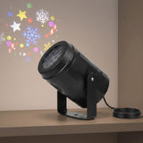 Black Christmas light projector sits tilted on a beige shelf beside coiled cable, projecting colorful stars, snowflakes, and candy canes onto the wall.