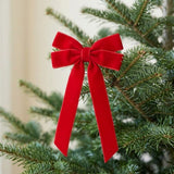 A red velvet bow with two long tails decorates a green Christmas tree branch against a softly blurred cream background.