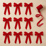 Ten red velvet Christmas bows with long tails lie on a pale wooden tabletop beside a spool of red ribbon.