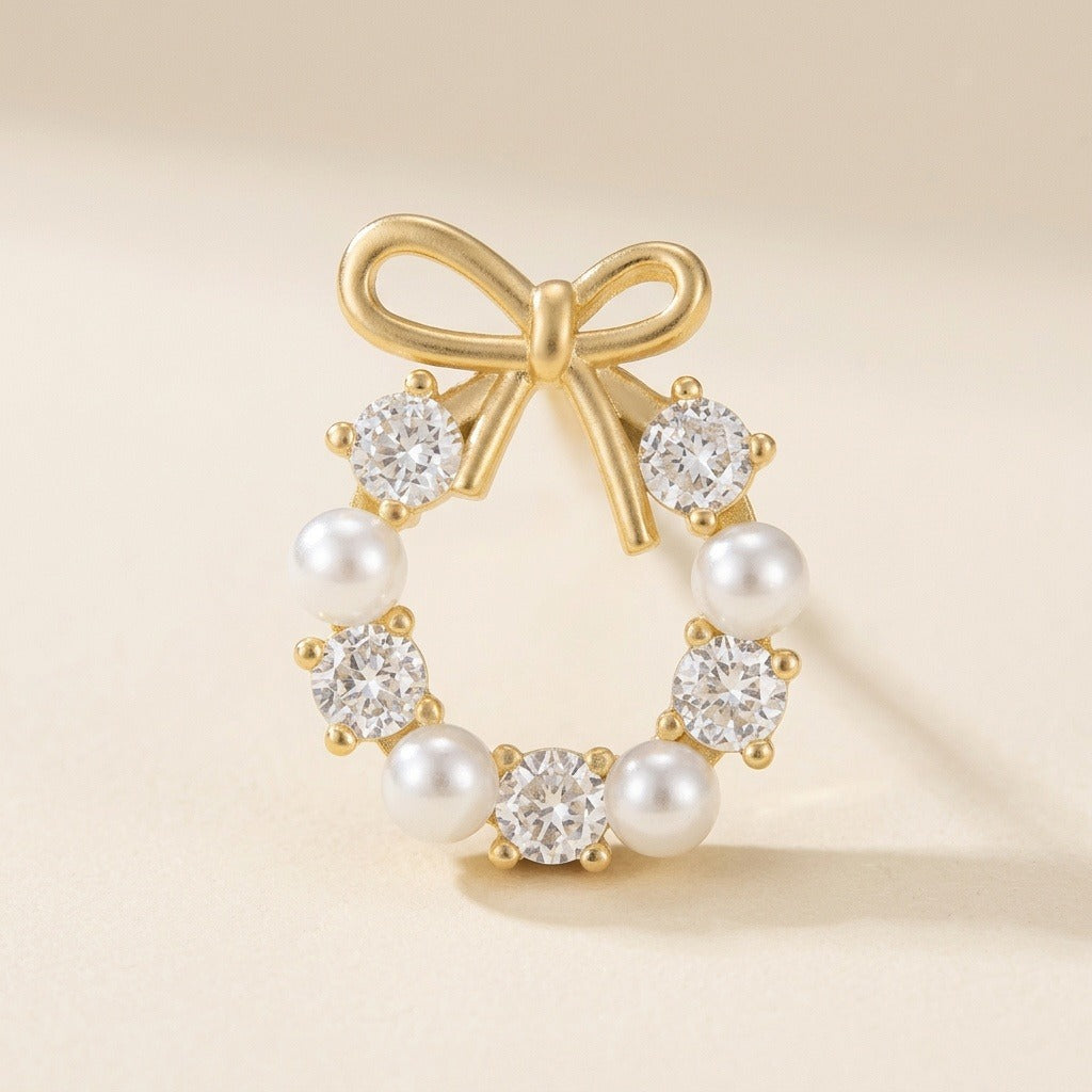 A gold-tone bow stud earring with white pearl-like beads and clear faceted stones stands on a cream surface, casting a shadow.