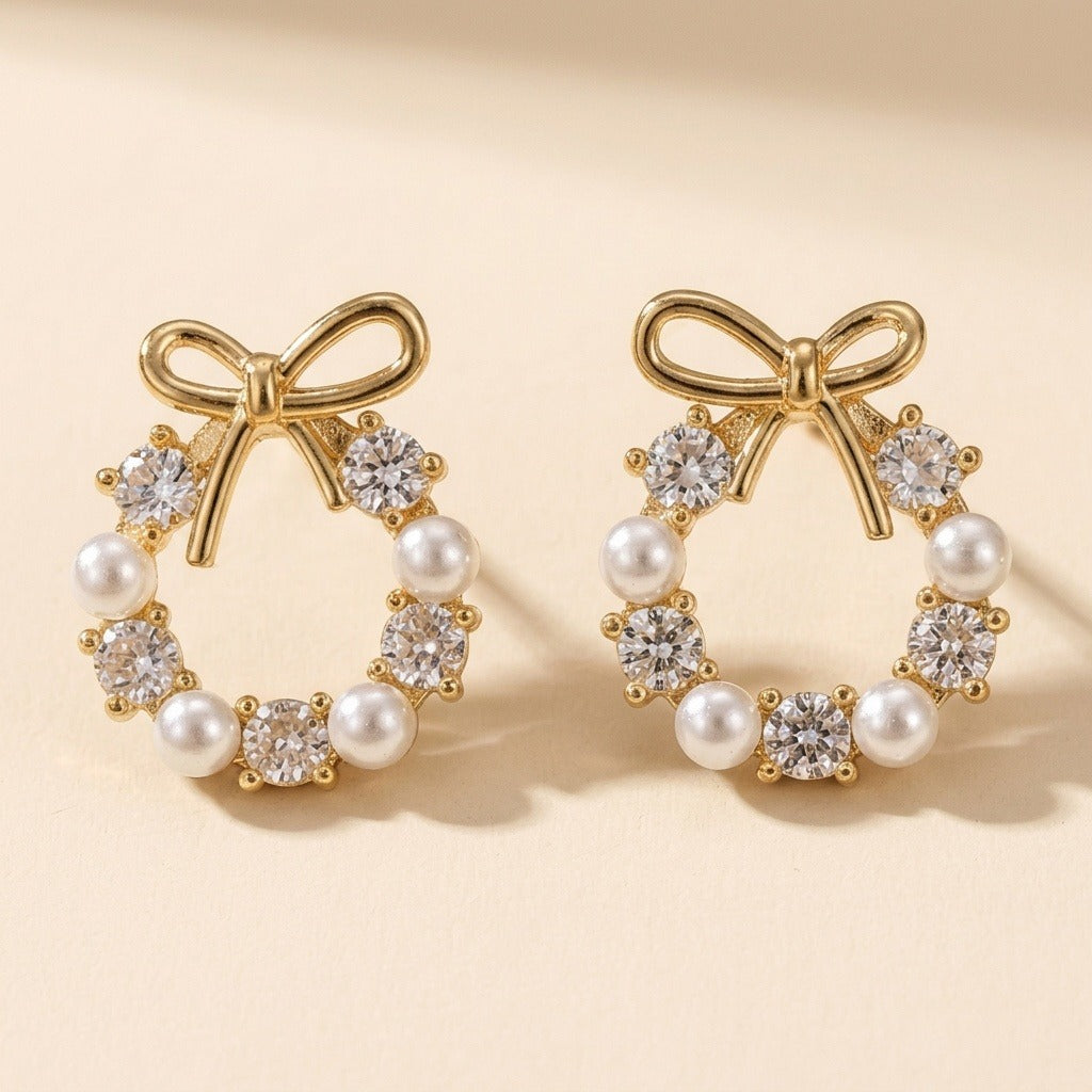 Stud earrings with gold-tone bows, white pearl-like beads and clear faceted stones stand against a warm cream background, casting shadows.
