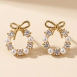 Stud earrings with gold-tone bows, white pearl-like beads and clear faceted stones stand against a warm cream background, casting shadows.