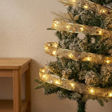 A glowing gold ribbon light string wraps around a snow-dusted Christmas tree beside a wooden table against a cream wall.