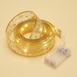 Coiled sheer gold ribbon light string with star patterns glows warm white beside a clear battery box on a cream background.